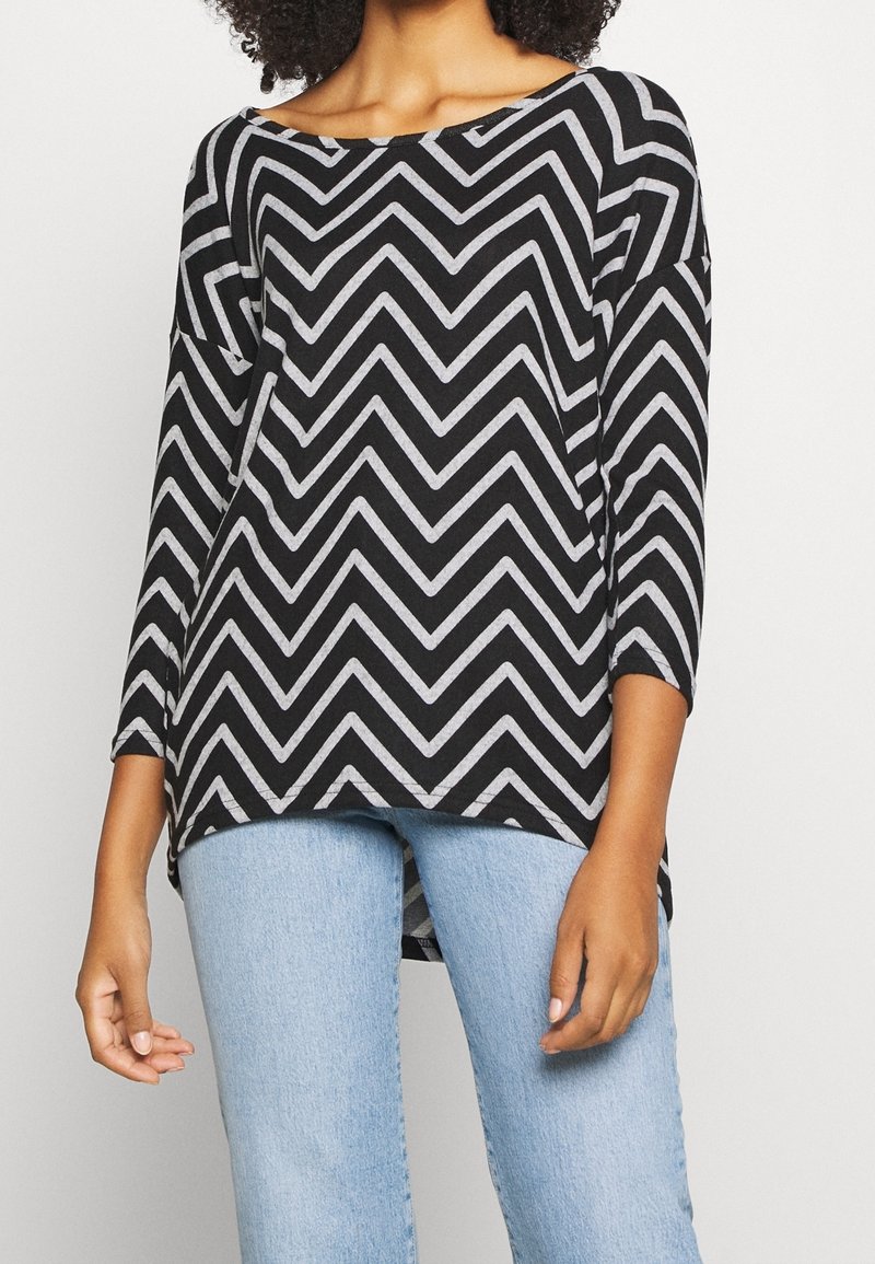 Musta ja harmaa chevron-kuvioinen yläosa, jossa on leveä kaula-aukko ja 3/4-hihat, yhdistettynä vaaleansinisiin farkkuihin. Materiaalin vaikuttaa olevan pehmeä ja kevyt.