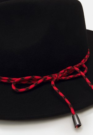 Chapeau noir à large bord avec un cordon rouge et noir à motifs noué en nœud autour de la base, se terminant par une pointe en métal argenté.