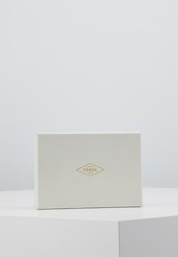 Witte gestructureerde cadeau-doos met een gouden logo waarop "FOSSIL" en "ISSUE No. 1954" op de voorkant staat, met een rechthoekige vorm en scherpe randen.
