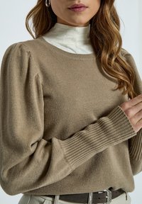 Bež pleteni pulover z rebrastimi manšetami, okroglo izreznico in napihnjenimi dolgimi rokavi, položen preko bele bluze z visokim ovratnikom in vzorcem mreže.