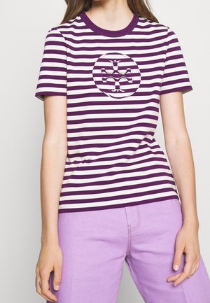 Frau trägt ein violett-weiß gestreiftes T-Shirt mit einem runden Logo und hellvioletten Hosen, steht vor einem einfarbigen Hintergrund.