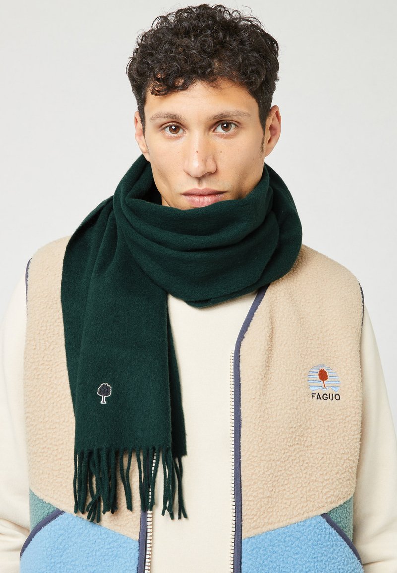 Groene wolschal met franjes aan de uiteinden, gedragen over een beige fleece jack met een kleurrijk geometrisch ontwerp en logodetails.