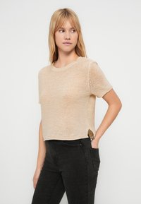 Bež kratki pleteni džemper s kratkim rukavima, rebrastim okovratnikom i bočnim prorezima. Nošen s tamnim trapericama, prikazujući opušteni kroj i teksturu.