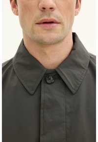 Mörkgrön regnjacka med vikbar krage, enkel knappstängning, slät textur och sydda sömmar, med en minimalistisk design.