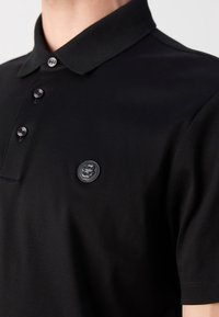 Polo noir avec un col classique, doté de trois boutons et d'un écusson logo texturé sur la poitrine gauche. Texture de tissu lisse.
