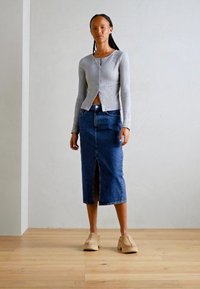 Cardigan a maniche lunghe grigio chiaro a coste, gonna in denim blu con spacco frontale, scarpe con piattaforma color beige, in piedi su un pavimento di legno contro una parete neutra.