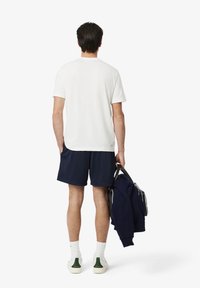 Camiseta blanca, pantalones cortos azul marino, llevando una chaqueta azul oscuro con detalles claros, calcetines blancos y calzado con ribetes verdes; visto desde atrás.