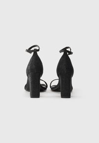 Sandali con tacco a blocco in glitter nero con cinturini alla caviglia. Presentano una texture liscia e un design open toe per favorire la ventilazione.