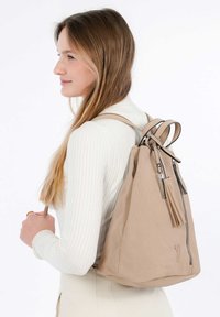 Beige perforerad läderryggsäck med justerbara remmar, dragkedja upptill och tofsdetalj. Mjuk textur och rundad form.