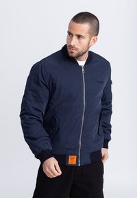 Marineblauwe bomberjack met ritssluiting, geribbelde manchetten en een oranje label op de zoom. Het materiaal lijkt glad en gewatteerd.