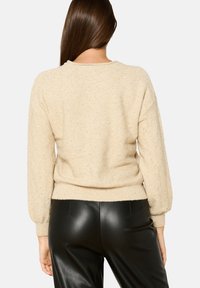 Beige gestrickter Pullover mit strukturiertem Finish, Rundhalsausschnitt und geripptem Saum; getragen mit schwarzen Lederhosen.