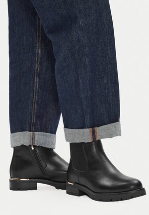 Czarne skórzane botki za kostkę z elastycznymi panelami po bokach, płaską podeszwą i złotym akcentem przy pięcie, noszone z podwiniętymi ciemnymi jeansami.