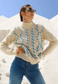 Frau mit zurückgekämmt Haar, die Sonnenbrillen, silberne Ohrringe, einen groben cremefarbenen Pullover mit blauen Akzenten und eine blaue Jeans trägt, vor einem schneebedeckten Hintergrund.
