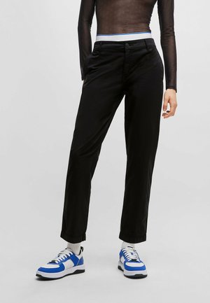 Person trägt schwarze Hose, weiße Socken und blaue und weiße Turnschuhe, steht vor einem einfarbigen hellen Hintergrund.