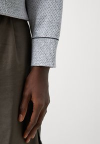 Ljusgrå texturerad tröja med ett stickat mönster och mörkblå accent vid muddarna, i kombination med en slät brun tyg under.