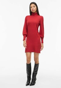 Vestido de punto rojo con un cuello alto, mangas largas abullonadas y una silueta ajustada. Combinado con botas negras de tacón alto hasta la rodilla.