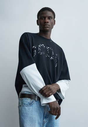 Νεαρός άνδρας που φοράει μαύρα και άσπρα πουκάμισα με στρώσεις και ανοιχτό μπλε τζιν, στέκεται μπροστά σε μια απλή ανοιχτογκρι φόντο.