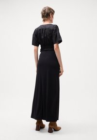 Robe maxi noire avec un buste smocké, des manches courtes bouffantes, un tissu lisse et une silhouette évasée. Associée à des bottes à talons hauts marron.