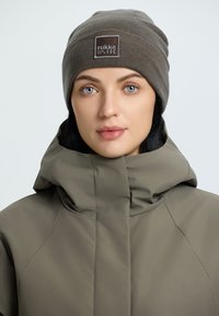 Ruskea pipo, jossa on tekstuuri neule ja pieni logo-tila. Oliivinvihreä takki korkealla kauluksella, sileästä kankaasta ja strukturoitu istuvuus.