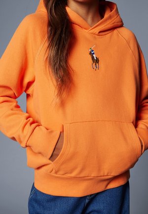 Person iført en orange hættetrøje med en frontlomme og en lille broderet polo-spiller logo på brystet, hænder i lommerne.