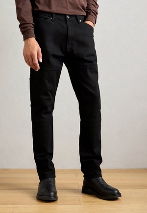 Man in strakke zwarte broek en zwarte leren laarzen staat op een houten vloer tegen een effen lichte muur.
