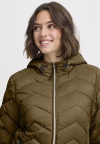 Chaki spalvos pūkinė striukė su chevron raštinimu, užtrauktuku užsegimu ir reguliuojamais raišteliais gaubte. Minkšta tekstūra ir lengvas dizainas.