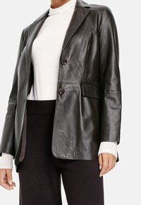 Czarna skórzana marynarka o gładkiej fakturze, jednorzędowym kroju, z klapami z wycięciem wcięcia oraz dwoma przednimi kieszeniami, noszona na białej bluzce.