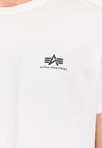 Balts kokvilnas T-krekls ar melnu logotipu, kurā attēlots stilizēts "A" burts ar spārniem kreisajā krūtīs, ar apaļu apkakli un īsām piedurknēm.