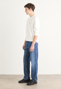 Camisa blanca de manga larga con cuello redondo, combinada con jeans de mezclilla azules y zapatillas negras, de pie sobre un suelo neutral contra un fondo claro.