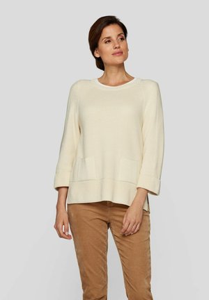 Cremfarbener Strickpullover mit rundem Ausschnitt, Dreiviertelärmeln, zwei vorderen Taschen und einer lässigen Passform, kombiniert mit beigen Hosen.