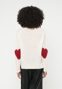 Krēmkrāsas džemperis ar faktūru pinumu un sarkaniem sirds akcentiem uz katra elkoņa, ar brīvu piegriezumu un ribotas aproces un apakšmalu.