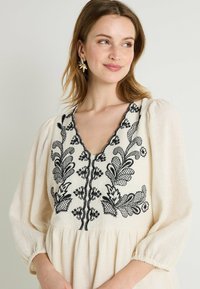 Vestido de color crema con bordados florales negros, escote en V profundo y mangas abullonadas, que presenta una cintura fruncida y una tela texturizada.