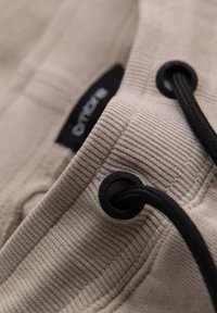 Gros plan sur un tissu beige avec une texture côtelée, un cordon noir et un œillet circulaire. Étiquette visible avec le nom de la marque en noir.