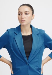 Modrý blazer se strukturovaným designem, klopami, jednobuttonovým zapínáním a bočními kapsami. Nosit na černém žebrovaném topu.