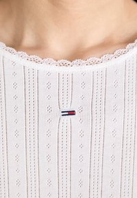 Biały sweter z dzianiny z delikatnym haftowanym wykończeniem, pionowymi żebrowaniami i małym, haftowanym logo z czerwonymi i niebieskimi akcentami na środku.