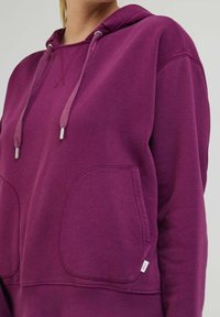 Lila huvtröja med snörning i huvan, kängurufickor och en mjuk textur. Har silverfärgade metallaccenter på snoddarna.