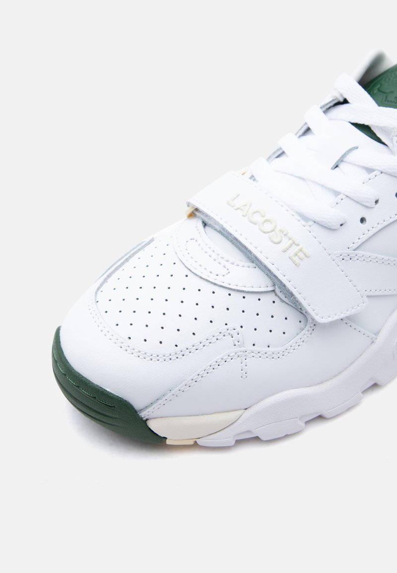 White Lacoste sneaker with perforated toe, white laces, green sole tip, and Velcro strap branded with "LACOSTE" in cream letters.