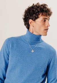 Blauer Rollkragenpullover aus weicher Wolle mit geripptem Ausschnitt. Trägt eine silberne, runde Anhänger-Kette.