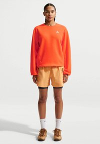 Γυναίκα που στέκεται φορώντας φωτεινή πορτοκαλί μπλούζα με μακριά μανίκια, ανοιχτό πορτοκαλί σορτς με μαύρα εσώρουχα, λευκές κάλτσες και καφέ παπούτσια πεζοπορίας σε λευκό φόντο.