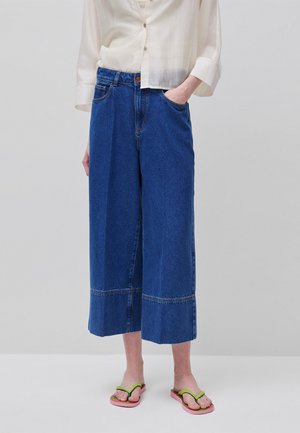 Persoon draagt een high-waisted, wijd uitgelegde cropped blauwe jeans, een crème geknoopt shirt en roze slippers met groene bandjes, staand tegen een effen achtergrond.