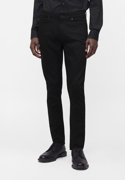 Uomo con pantaloni neri slim, camicia nera e scarpe eleganti nere in pelle con lacci, in piedi davanti a uno sfondo semplice.