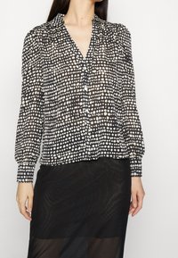 Black and white polka dot blouse with a v-neckline, long sleeves, button closure, and gathered detailing at shoulders. Light, semi-sheer fabric.