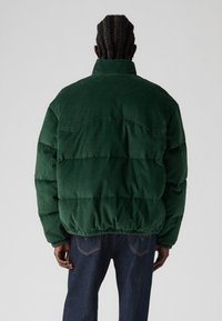 Zielona kurtka pikowana z sztruksu ma wysoki kołnierz, elastyczny dół i pikowany wzór. Z tyłu znajduje się karczek.