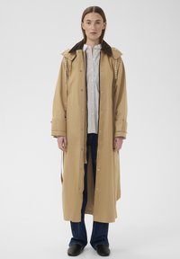 Žena stojí čelem vpřed, má na sobě dlouhý béžový trenčkot přes bílou košili, tmavé džínsy a černé boty na jednoduchém pozadí.