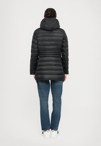 Zwarte pufferjas met een capuchon, met een gestikte textuur en slim fit. Gecombineerd met blauwe spijkerbroeken en witte sneakers, gezien van achteren.