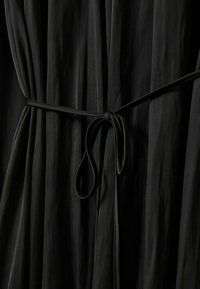 Juoda plisuota suknelė su surištu diržu, parodanti lygią tekstūrą ir tekantį dizainą. Audinys yra lengvas ir minkštas.