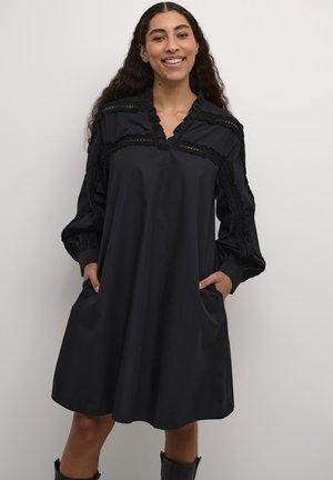 Black cotton dress, knee-length with a ruffled collar, lace accents, long sleeves, and two side pockets. Smooth texture with a loose fit.