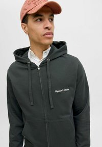 Mørkegrøn lynlås hoodie med snore og frontlommer, med hvidt broderet logo "Originals Studio" på brystet.