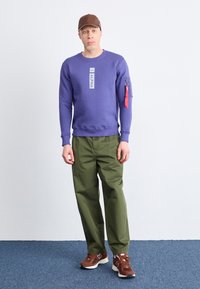Felpa viola con etichetta "ALPHA", dotata di tasca con zip sulla manica e accenti rossi. I pantaloni verde oliva e le sneaker marroni completano il look.
