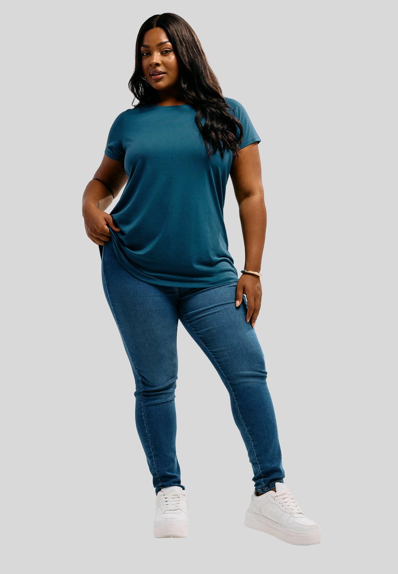 Donna con una maglietta a maniche corte color turchese, jeans blu e sneakers bianche, in piedi con una mano che tira la maglietta e l'altra poggiata sulla coscia.